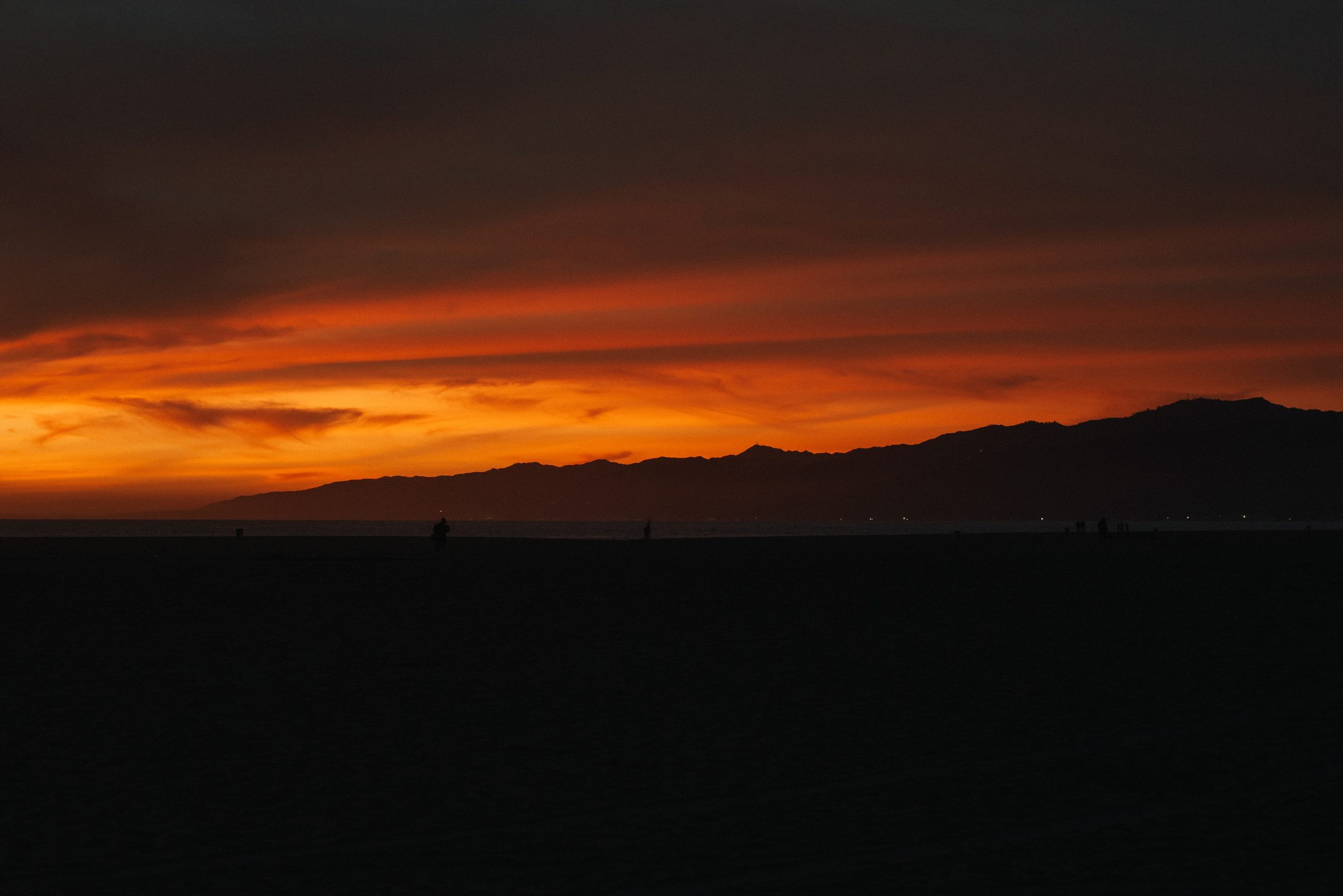 A dark beach under a band of deep orange sunset cloud, a mountain ridge in silhouette on the horizon.
