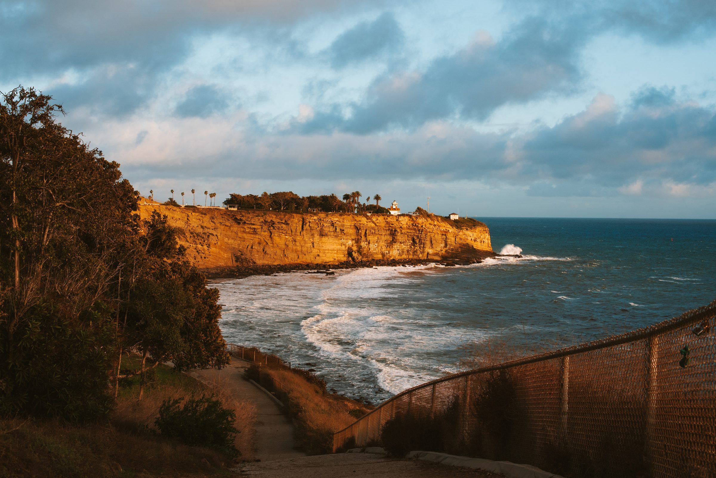 A sunlit golden sea cliff with a lighthouse on its point, white surf breaking below and a fenced path in the foreground.
