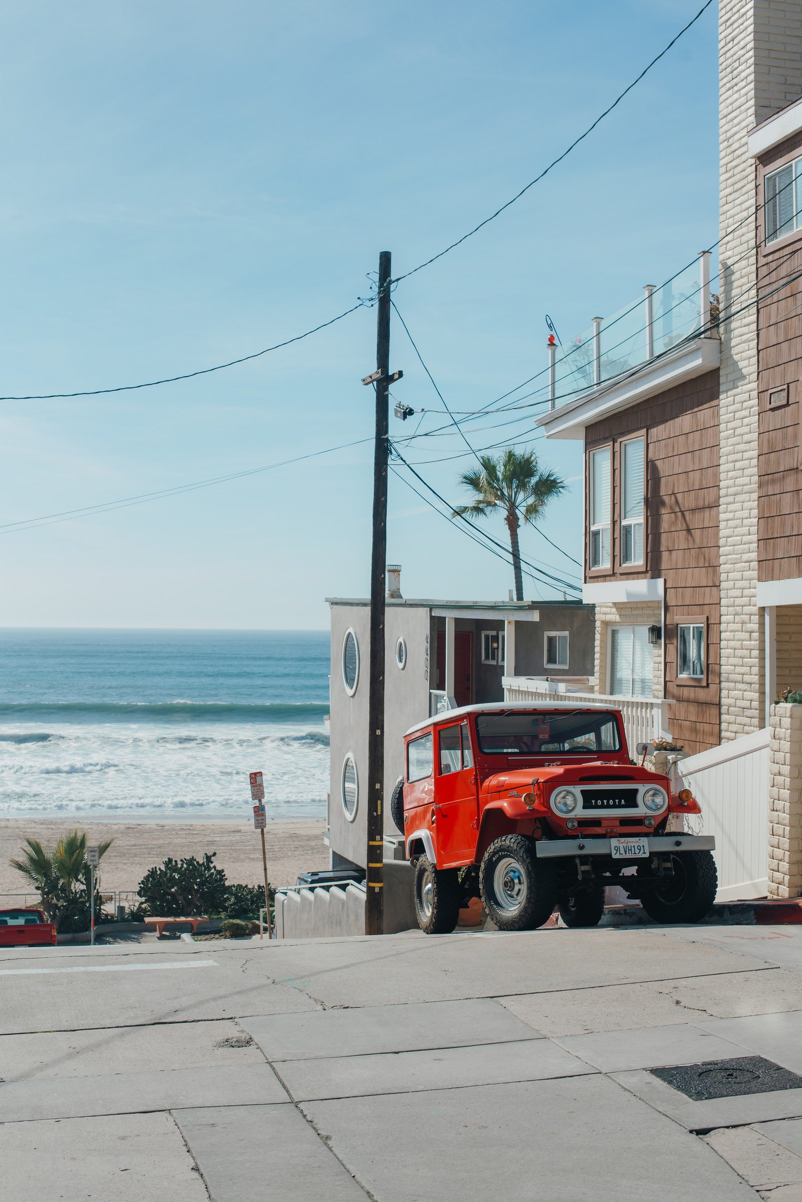 A red vintage Toyota Land Cruiser parked on a steep beach street beside a shingled house, with the ocean and a palm tree behind it.