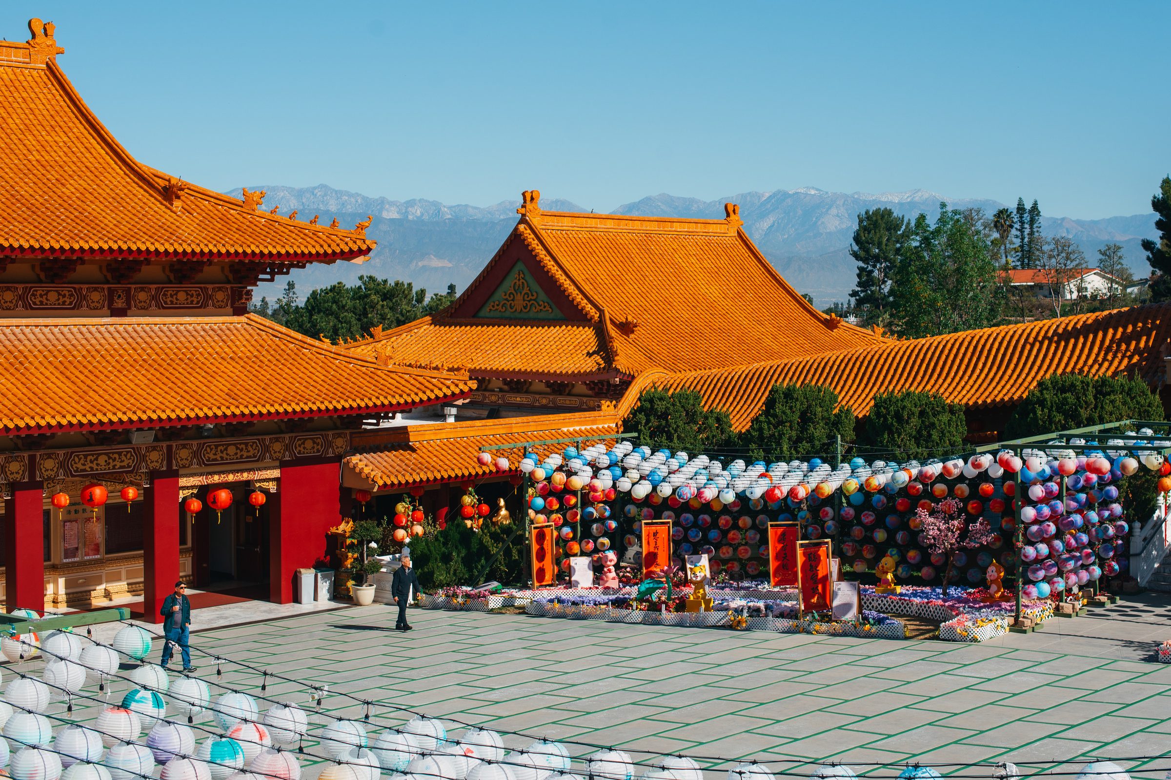 A Chinese temple courtyard with orange tiled roofs, red pillars and rows of colorful paper lanterns, mountains beyond.