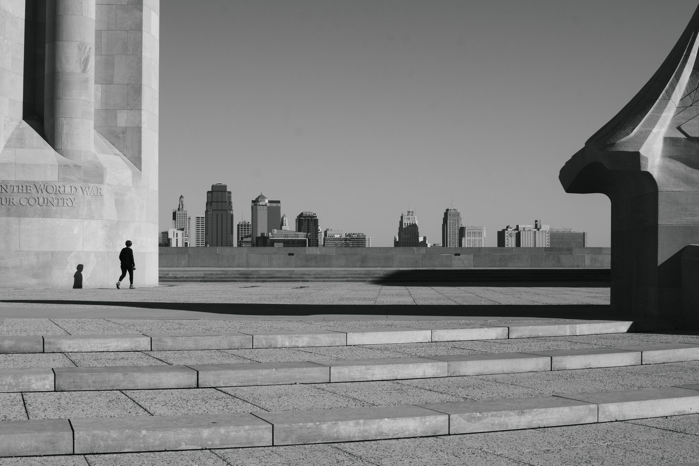 Black-and-white view across the stone plaza of a war memorial: a lone walker and their shadow at the base of a carved column, the Kansas City skyline low on the horizon, and broad curved steps in the foreground.