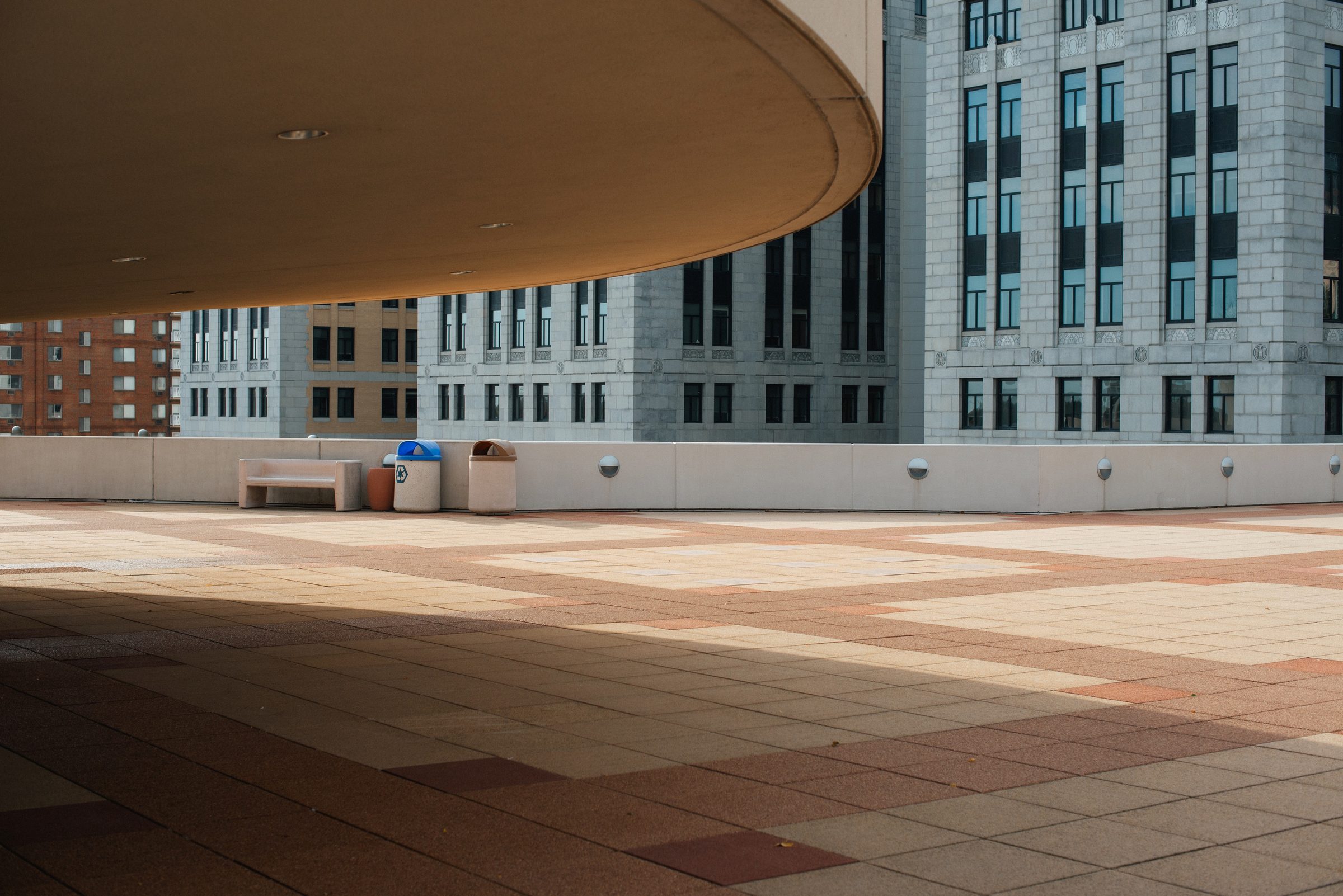 The curved underside of a concrete overhang above an empty tiled plaza, with tall gray downtown buildings beyond it.