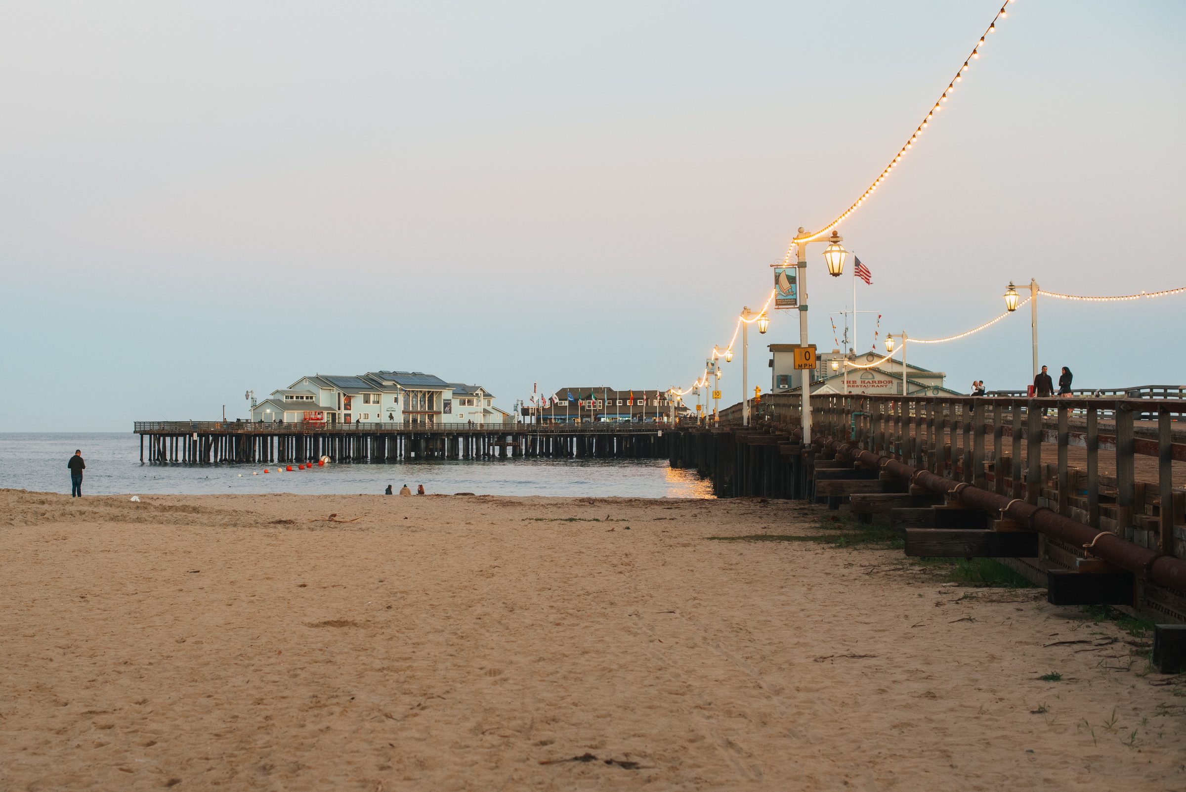 A long wooden pier strung with lights at dusk, a few people on the sand below and on the boardwalk.