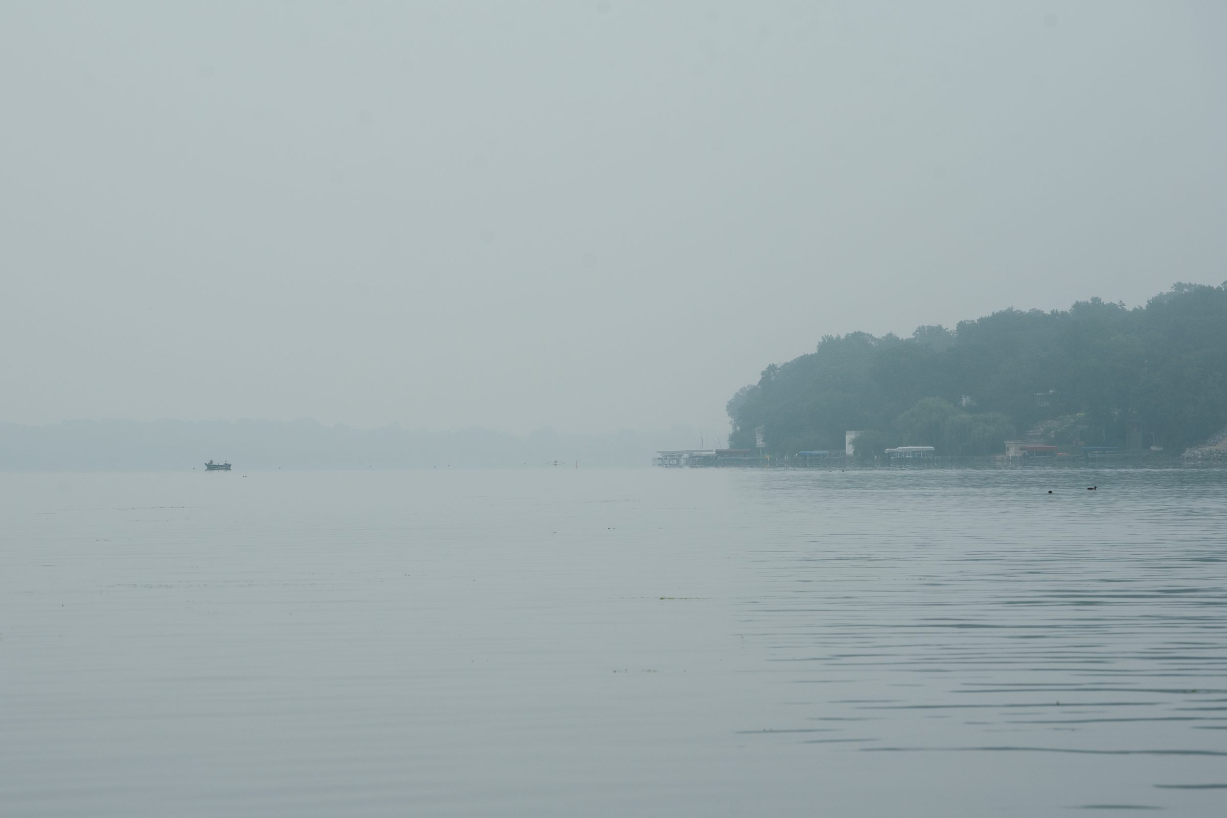 Lake Monona in heavy morning mist, the water flat and gray-green, a wooded point fading on the right and a small boat barely visible in the distance.
