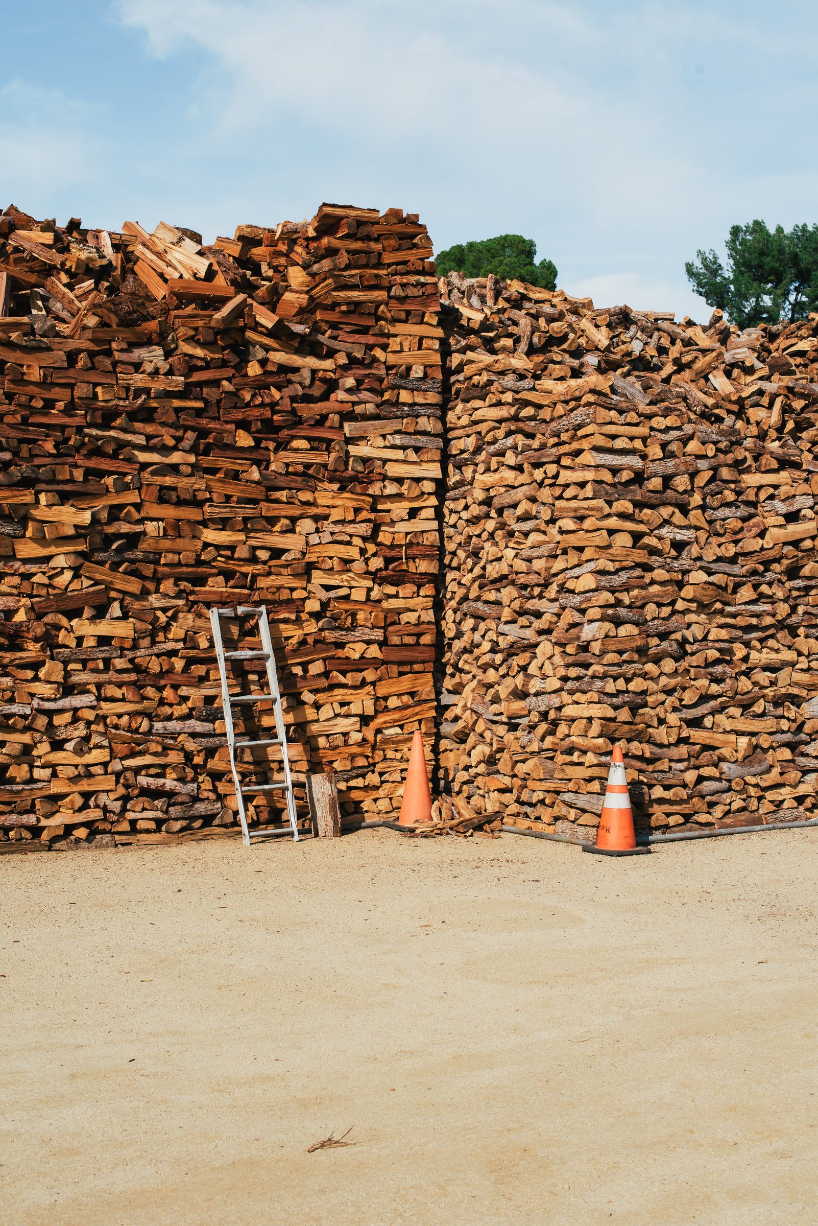A tall wall of stacked split firewood under a blue sky, with an aluminum ladder leaning against it and two orange traffic cones on the sandy ground below.
