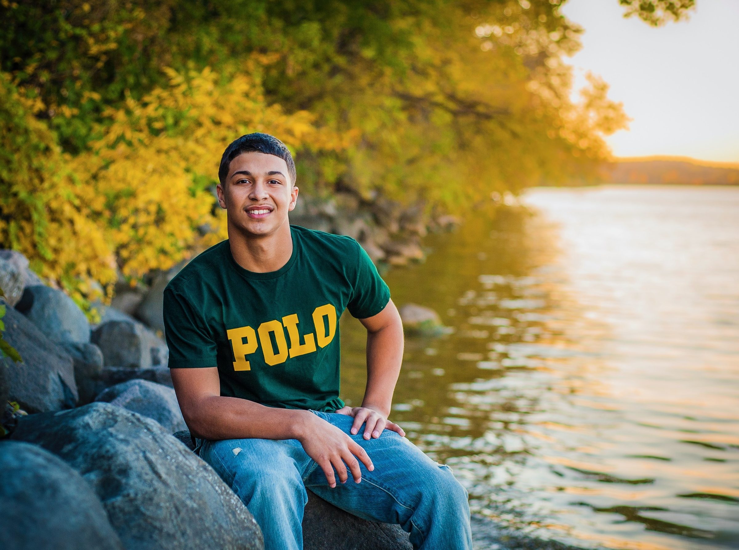 A young man in a green shirt sitting on lakeshore rocks in golden evening light.