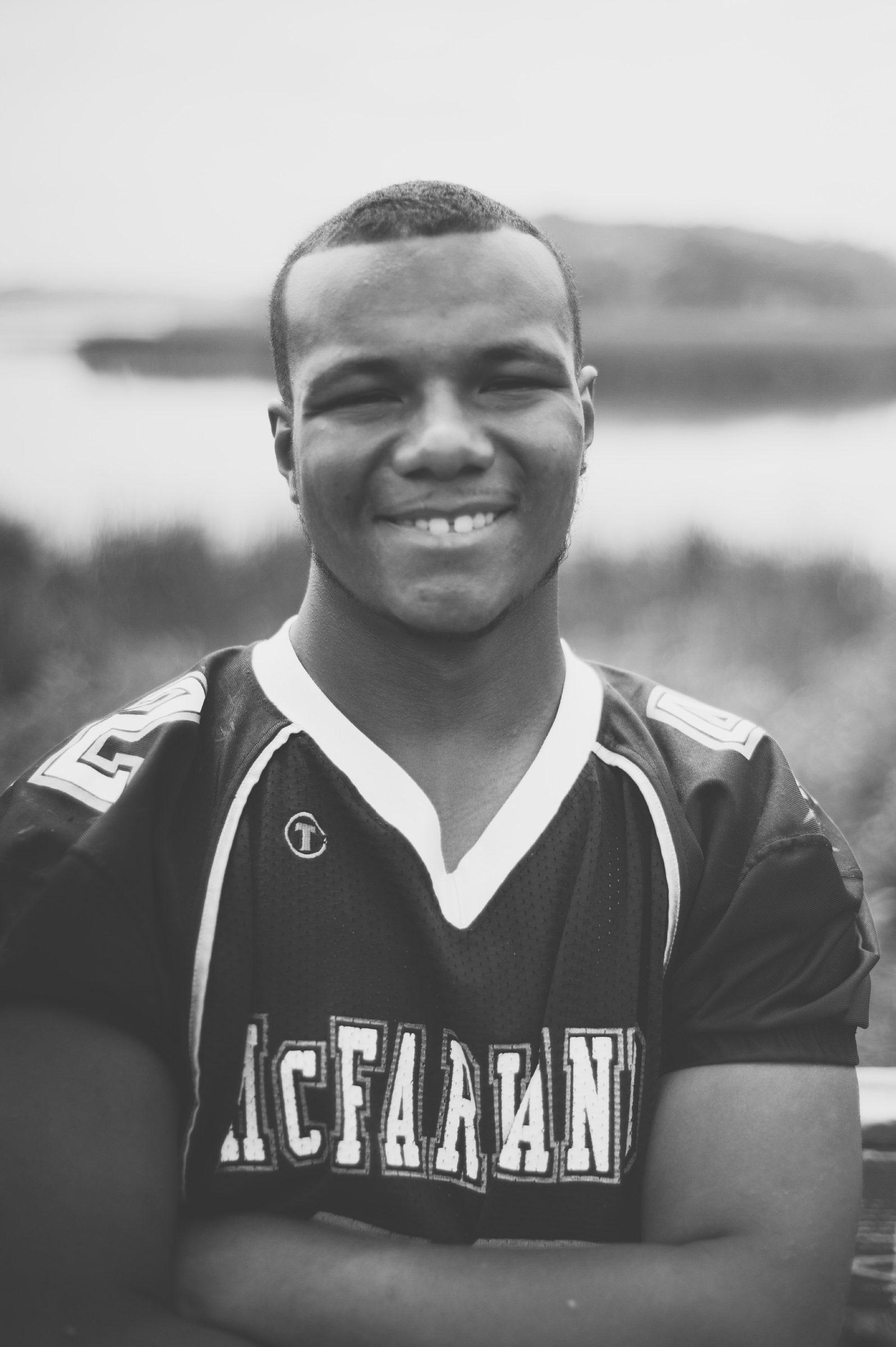 Black-and-white portrait of a smiling young man in a soccer jersey, a lake soft behind him.