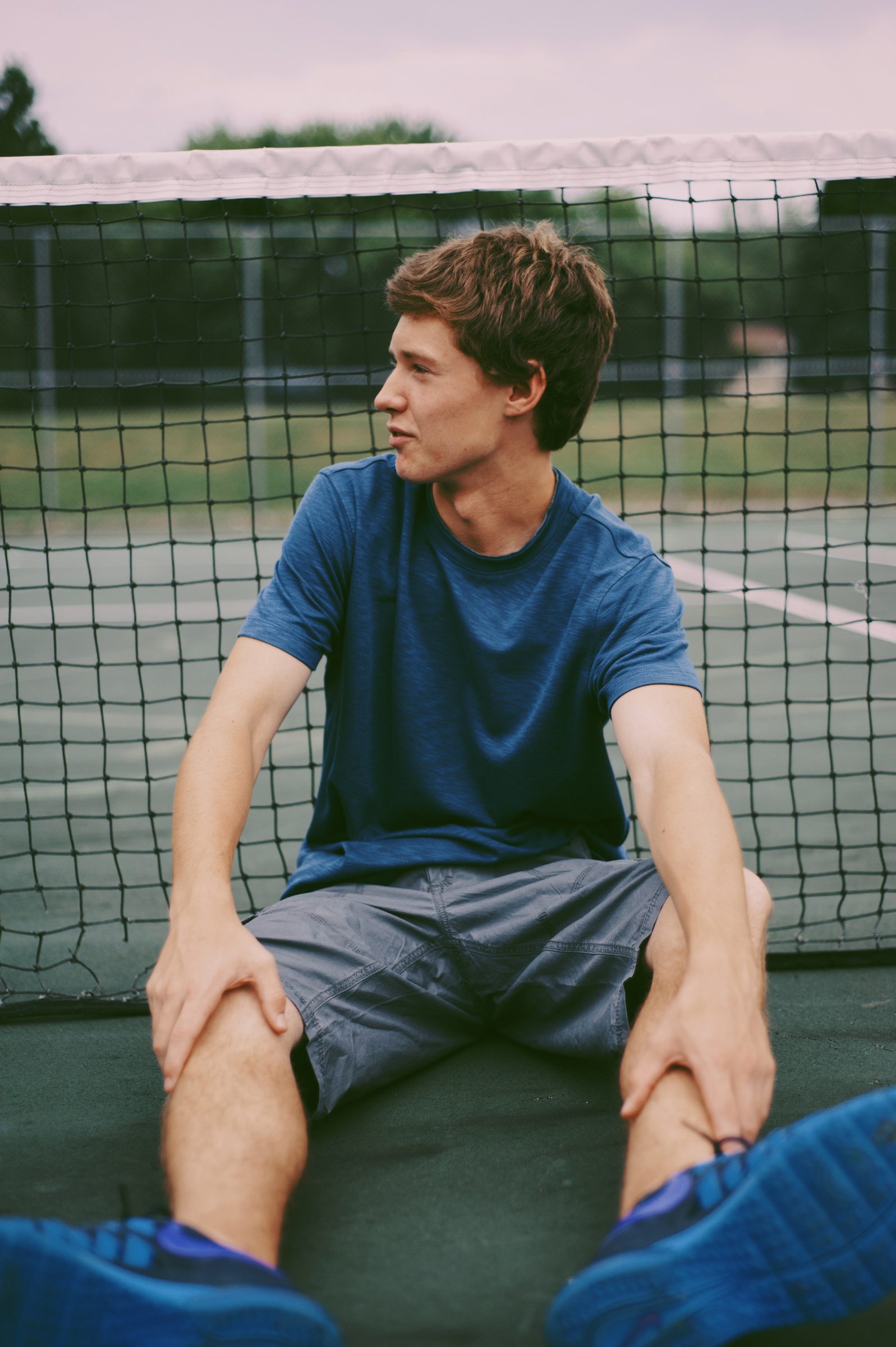 A young man in a blue T-shirt sitting against the net of a tennis court.
