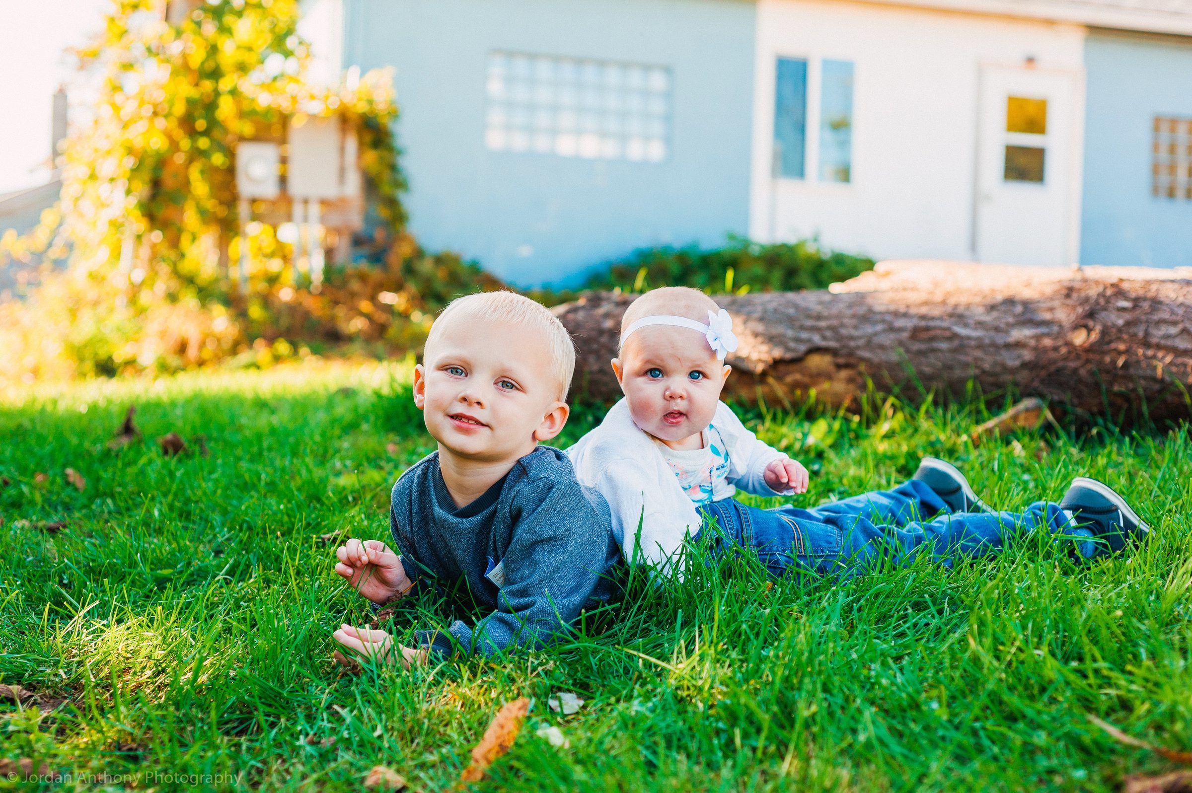 Two small children lying on the grass in front of a pale blue building.