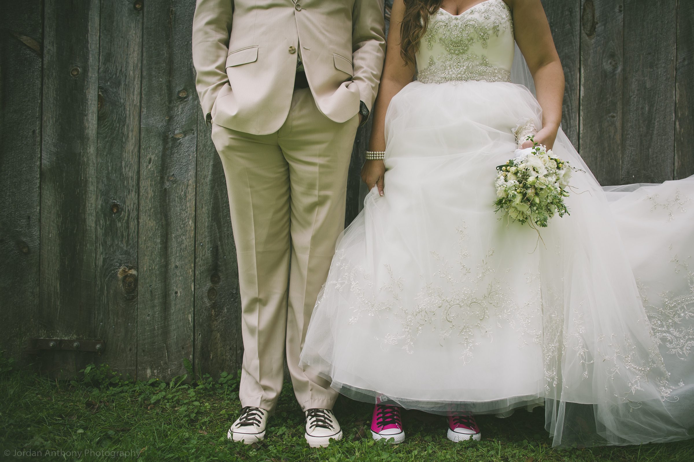Close detail of a bride and groom side by side, both in sneakers, against a weathered wooden fence.