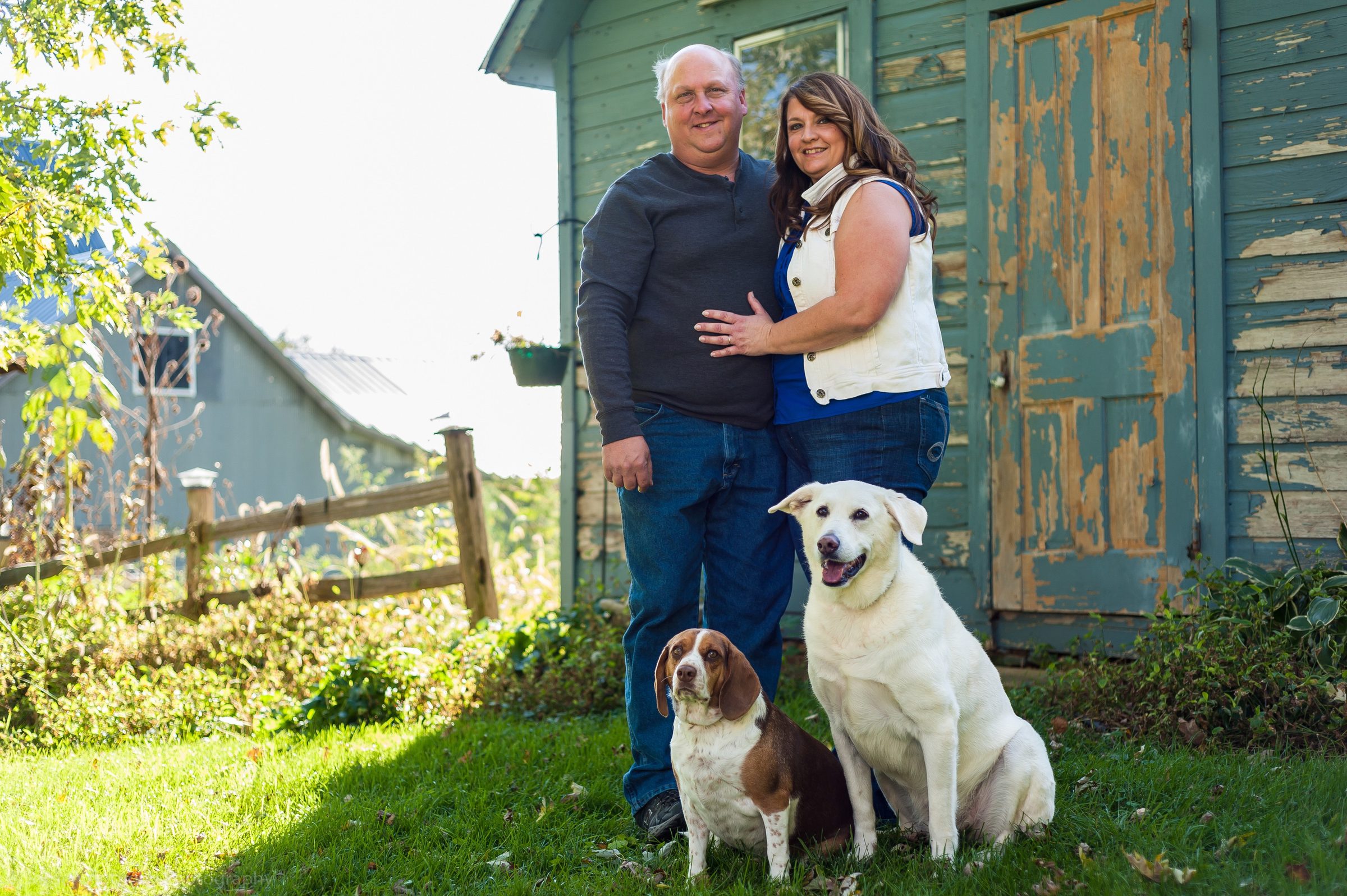 A couple standing with their two dogs in front of a weathered blue barn.
